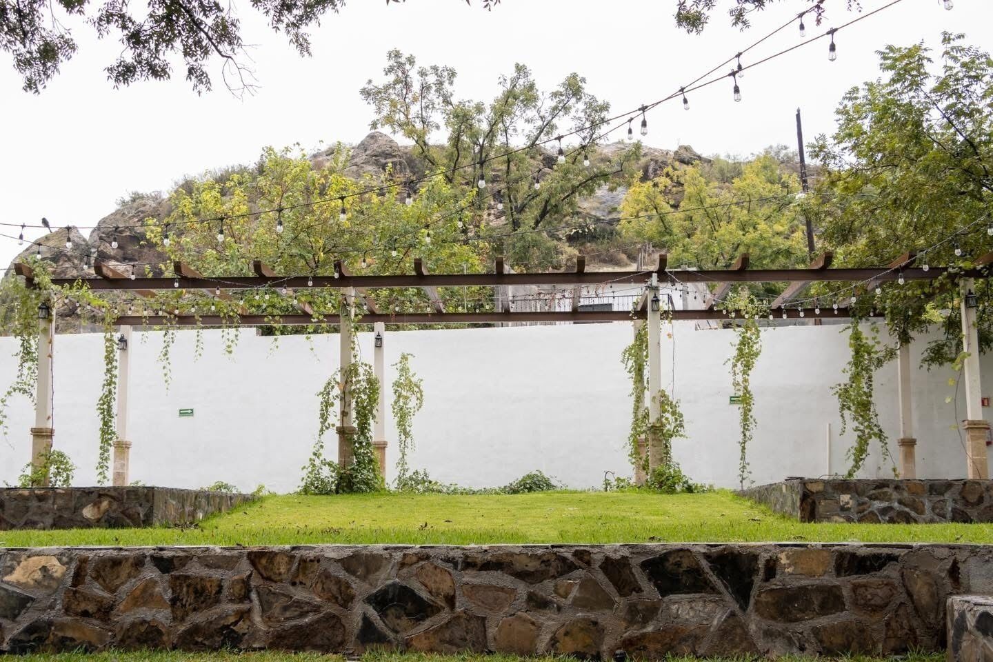 Una pérgola con luces de cuerda, un césped y un muro de piedra.