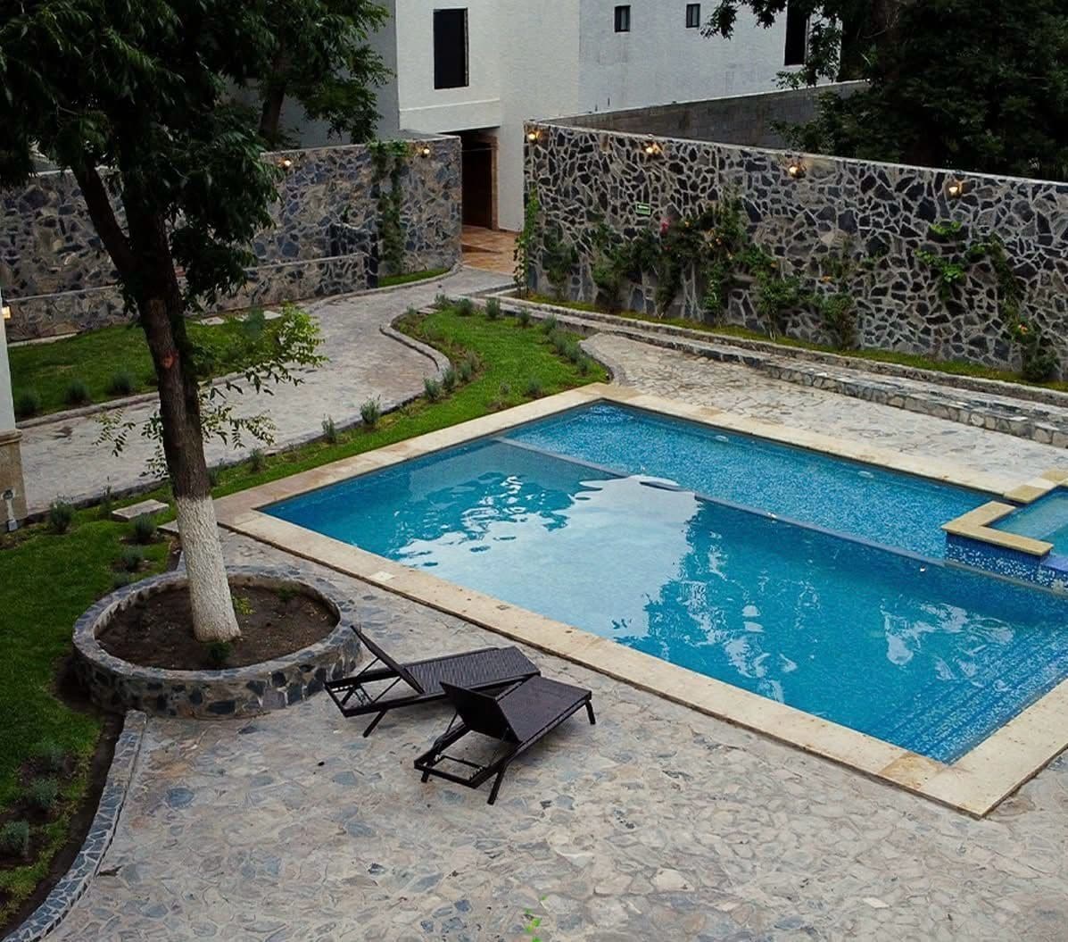 A rectangular swimming pool with a stone border, surrounded by a stone wall and a small garden area with a tree and two lounge chairs.