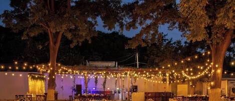 An outdoor event space with tables set up for dining, surrounded by trees and string lights.