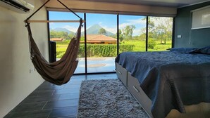 A bedroom with a bed, a hammock, a large window, and a view of a mountain and greenery.