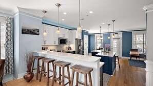 Una cocina moderna con una isla central, taburetes de bar, iluminación colgante y vistas al área de comedor.
