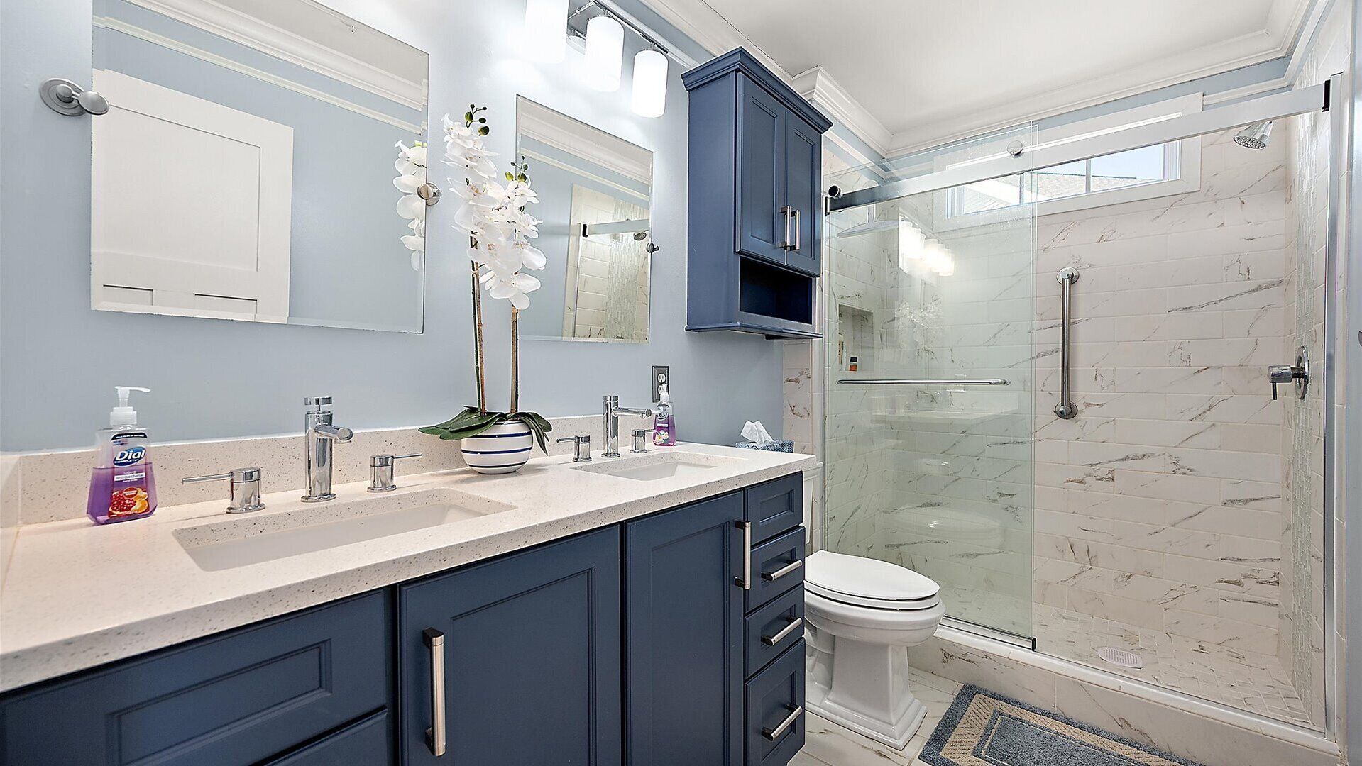 A bathroom with a double sink vanity, a toilet, a glass shower enclosure, and a marble-tiled shower area.