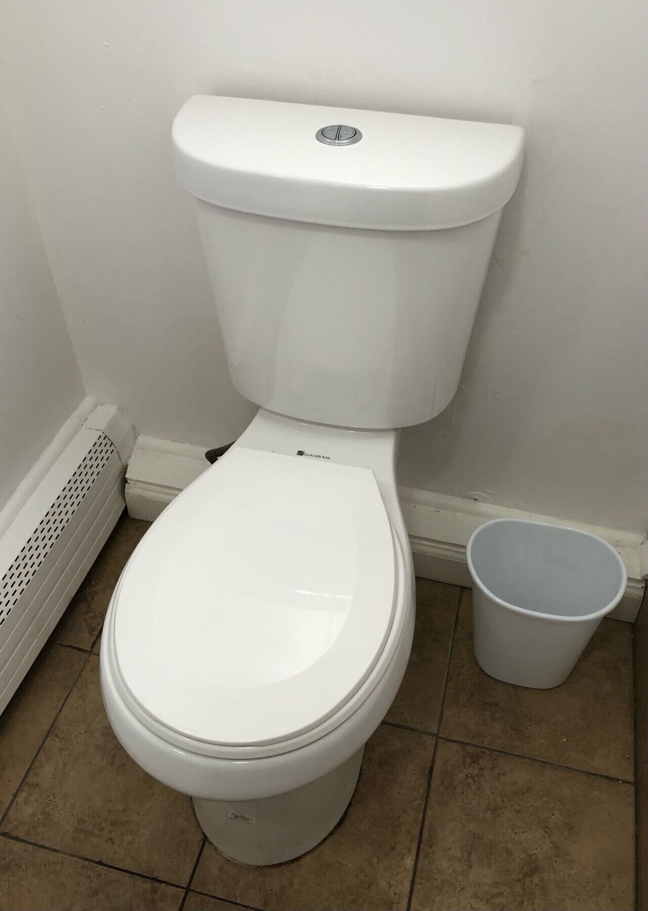 A white toilet with a closed lid, a white bucket next to it, and a vent on the left.