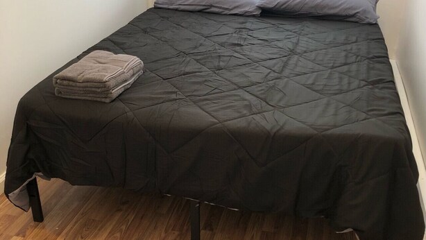 A single bed with a black quilted bedspread and two pillows, a folded towel on the bed, and a window above.