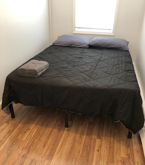 Una cama individual con un edredón acolchado negro y dos almohadas, una toalla doblada sobre la cama, y una ventana arriba.