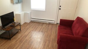 Una habitación con piso de madera, un sofá rojo, un televisor sobre un mueble bajo y una puerta blanca.