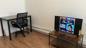Una habitación con piso de madera, una silla de oficina negra, una pequeña mesa de madera con un televisor y un escritorio negro.