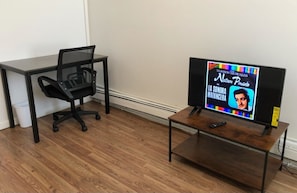 Una habitació amb terra de fusta, una cadira d'oficina negra, una taula de fusta petita amb un televisor i un escriptori negre.