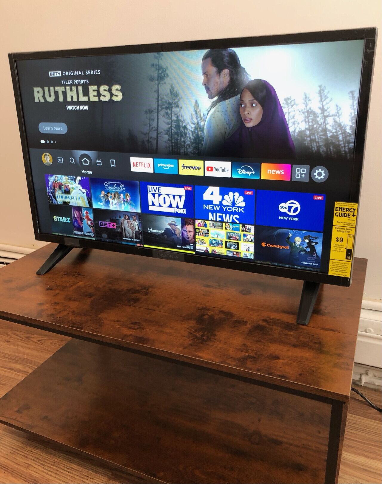 Televisor de pantalla plana sobre un soporte de madera que muestra la interfaz de una plataforma de streaming con varios canales y opciones de contenido.