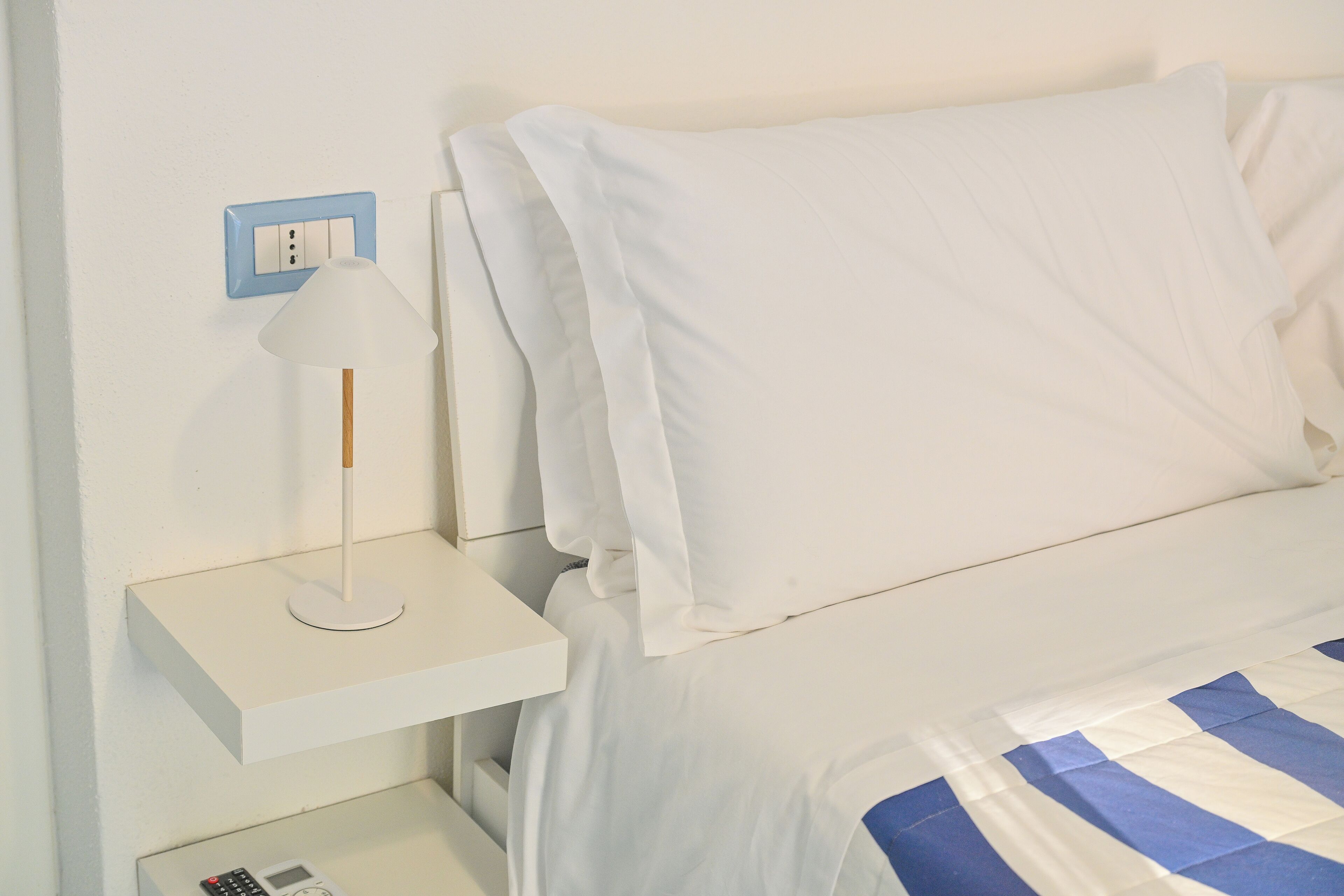 Una cama con ropa de cama blanca y una manta a rayas azul y blanco, una mesita de noche blanca con una lámpara, y un enchufe en la pared.