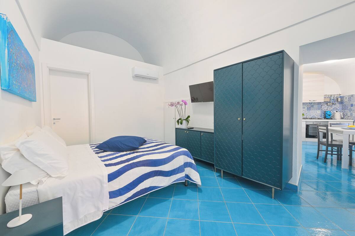 A bedroom with a bed, a wardrobe, a desk, and a small kitchen area.