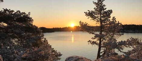 A serene lakeside scene with a clear view of the sunset over the water, surrounded by snow-covered trees.