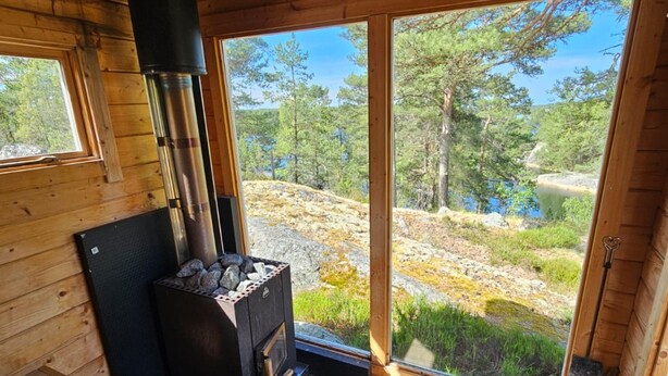 A wooden cabin interior with a wood-burning stove, large windows, and a view of a forest and lake. - Dream Cottage in the Stockholm Archipelago (Djurhamn)