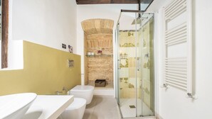 Une salle de bain avec un lavabo, des toilettes et une douche, le tout de couleur blanche. On y trouve un mur en pierre et une paroi de douche en verre.