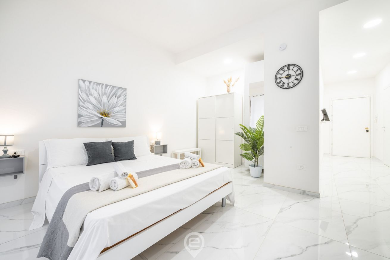A modern bedroom with a large bed, a bedside table with a lamp, a wall clock, and a potted plant.
