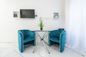 Una habitación moderna con una pared blanca, un televisor de pantalla plana montado sobre una mesita con una planta en maceta, y dos sillas color turquesa.