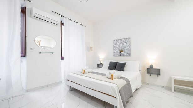 A modern bedroom with a large bed, a bedside table, a wall-mounted mirror, and a ceiling light fixture. - Guest Room Historic Centre (Iglesias)