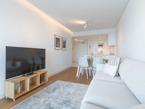 Ein modernes Wohnzimmer mit Flachbildfernseher, einem Holz-TV-Schrank, einem weißen Sofa und einem Essbereich mit Glastisch und Stühlen.