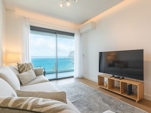 A modern living room with a large flat-screen TV, a sofa set, and a view of the ocean through a sliding glass door. - Glamorous Apartment Funchal by Atlantic Holiday (Funchal)