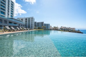 A modern hotel with a large infinity pool, lounge chairs, and a view of the ocean.