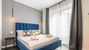 A modern bedroom with a blue headboard, a small bedside table, a neatly made bed with pillows and towels, and a large window with sheer curtains. - Saint Margaret Apartments - Olivia (Cagliari)