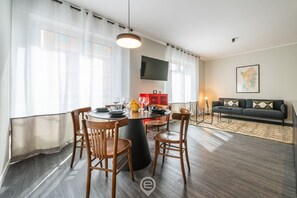 A modern living room with a round dining table, wooden chairs, a sofa, and a flat-screen TV mounted on the wall.