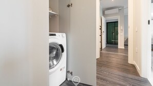 Une buanderie avec un lave-linge blanc, une porte donnant sur un couloir au plancher en bois, et une porte verte à l’arrière-plan.
