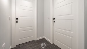 Un couloir avec deux portes blanches, chacune dotée d’une poignée noire et d’un trou de serrure. Le sol est en bois et les murs sont peints en blanc.