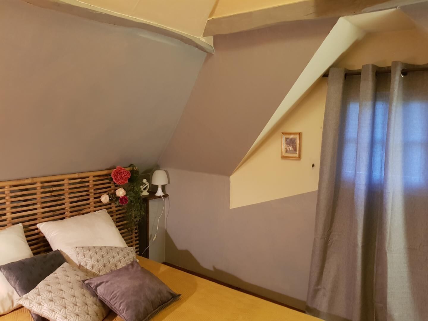 Um quarto com teto inclinado, uma cama com várias almofadas, uma luminária de cabeceira e uma janela com cortinas.