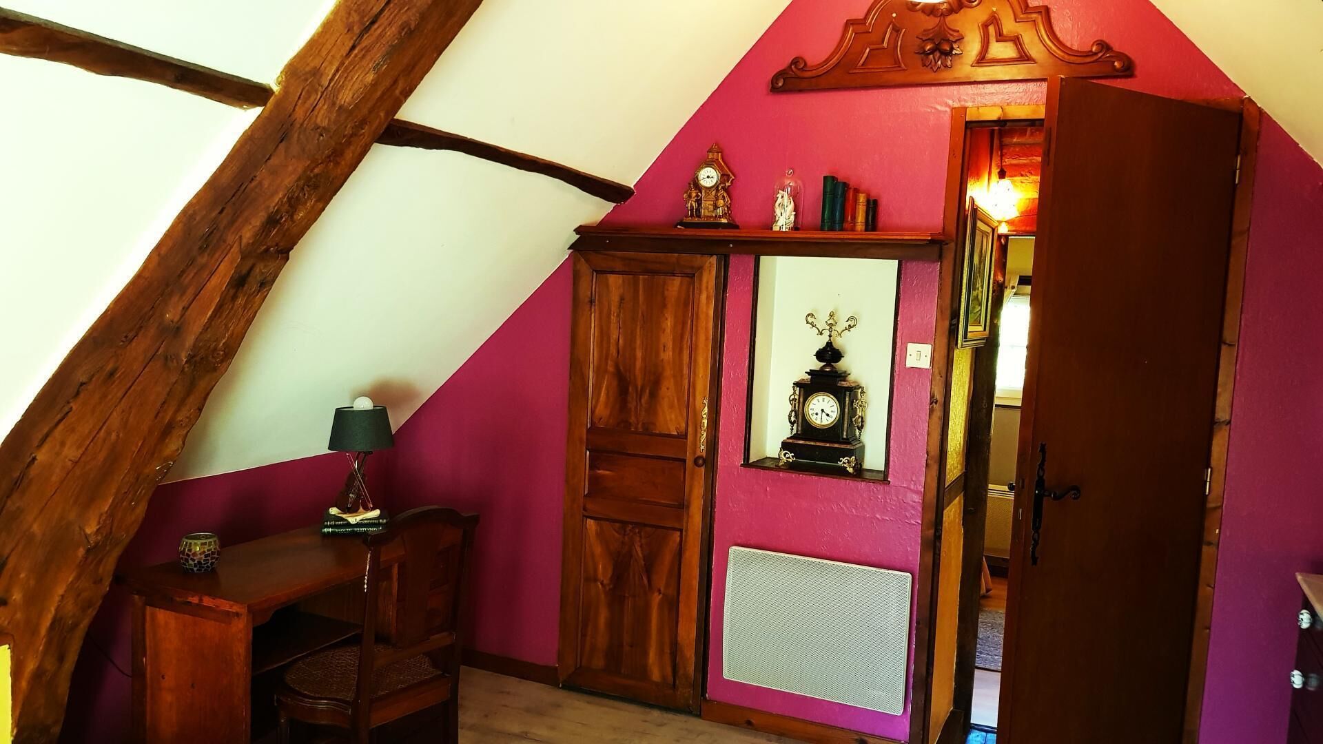 Ein Zimmer mit schrägverlaufender Decke, Holzbalken, einem Schreibtisch mit Lampe, einem Kleiderschrank, einem Spiegel und einem Heizkörper.