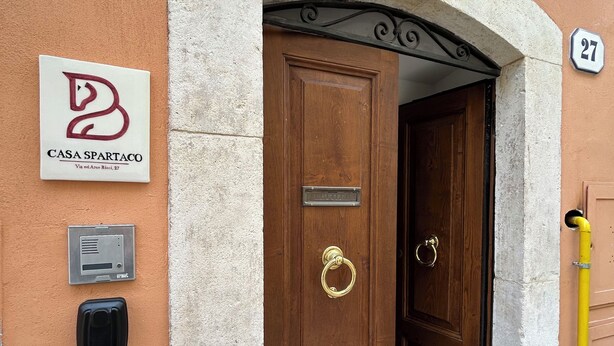 Une porte en bois ornée d’un heurtoir en laiton, une boîte aux lettres et une enseigne indiquant « Casa Spartaco » sur le mur.