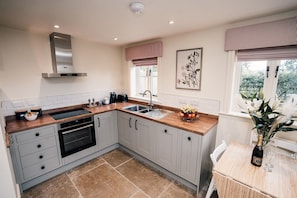A modern kitchen with grey cabinets, a wooden countertop, a stainless steel range hood, a built-in oven, a sink, and a window with a view of greenery.