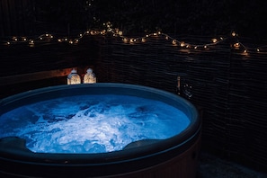 A hot tub with a wooden edge, surrounded by a stone wall, and illuminated by string lights.