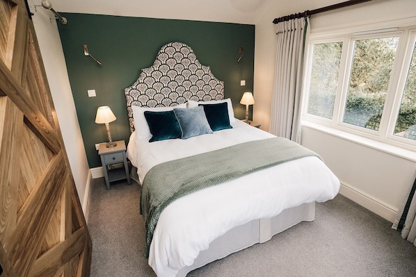 A neatly made bed with a patterned headboard, two bedside lamps, and a view of greenery through a window.
