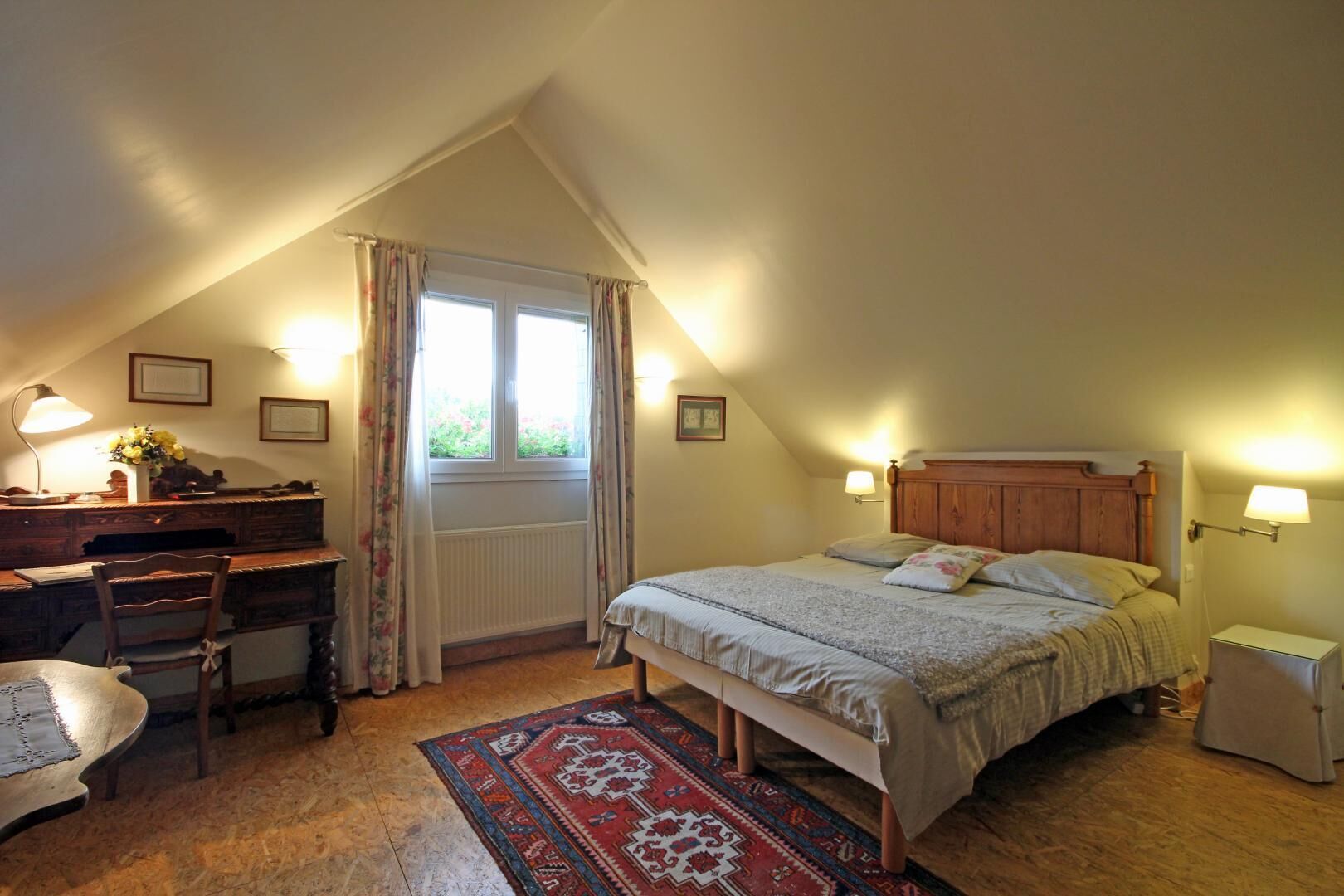 Un dormitorio acogedor con techo inclinado, un escritorio de madera, cama con cabecera y ventana con cortinas.
