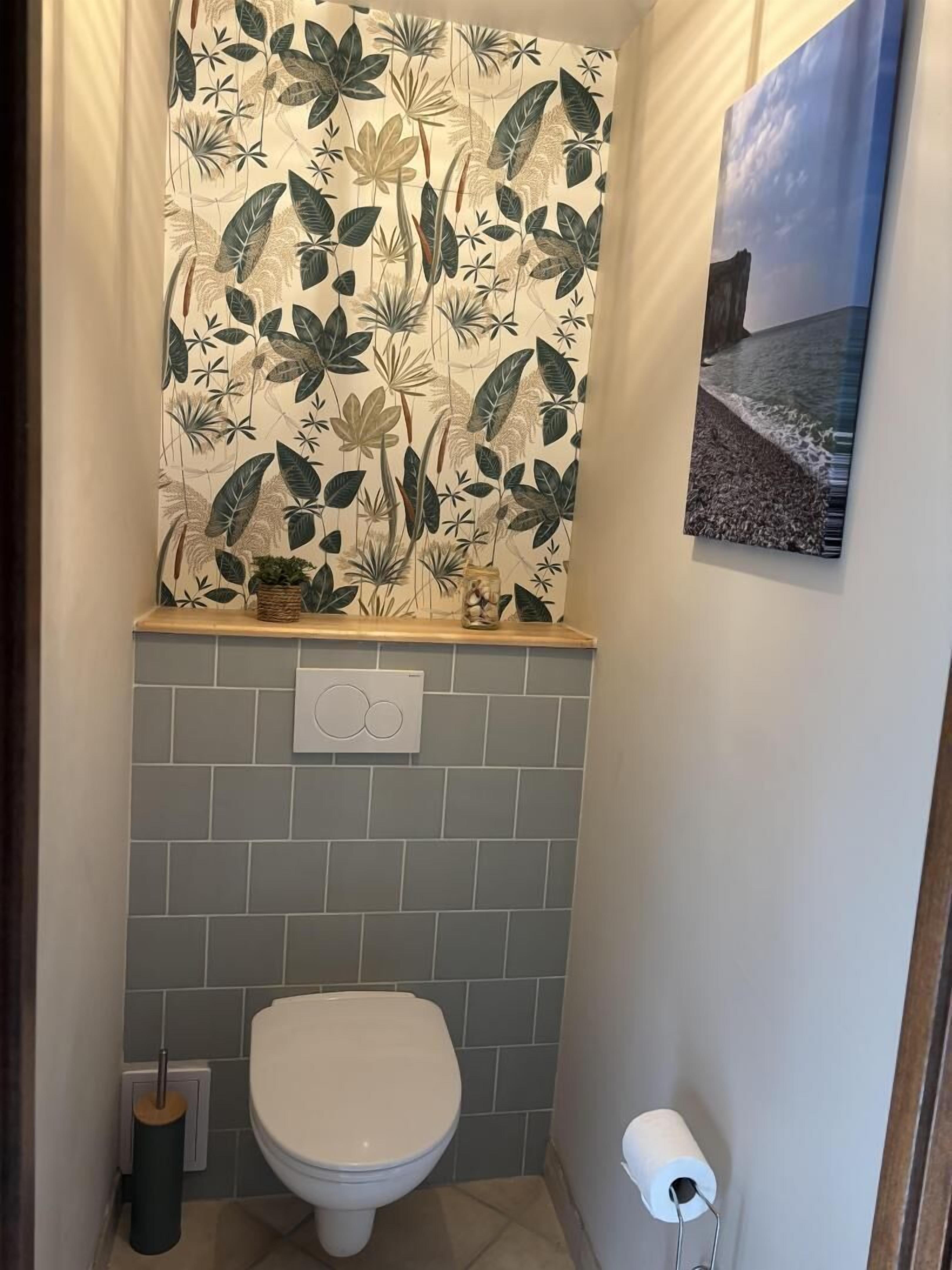 A bathroom with a toilet, a wall-mounted toilet paper holder, a tiled backsplash, and a shower curtain with a tropical leaf pattern.