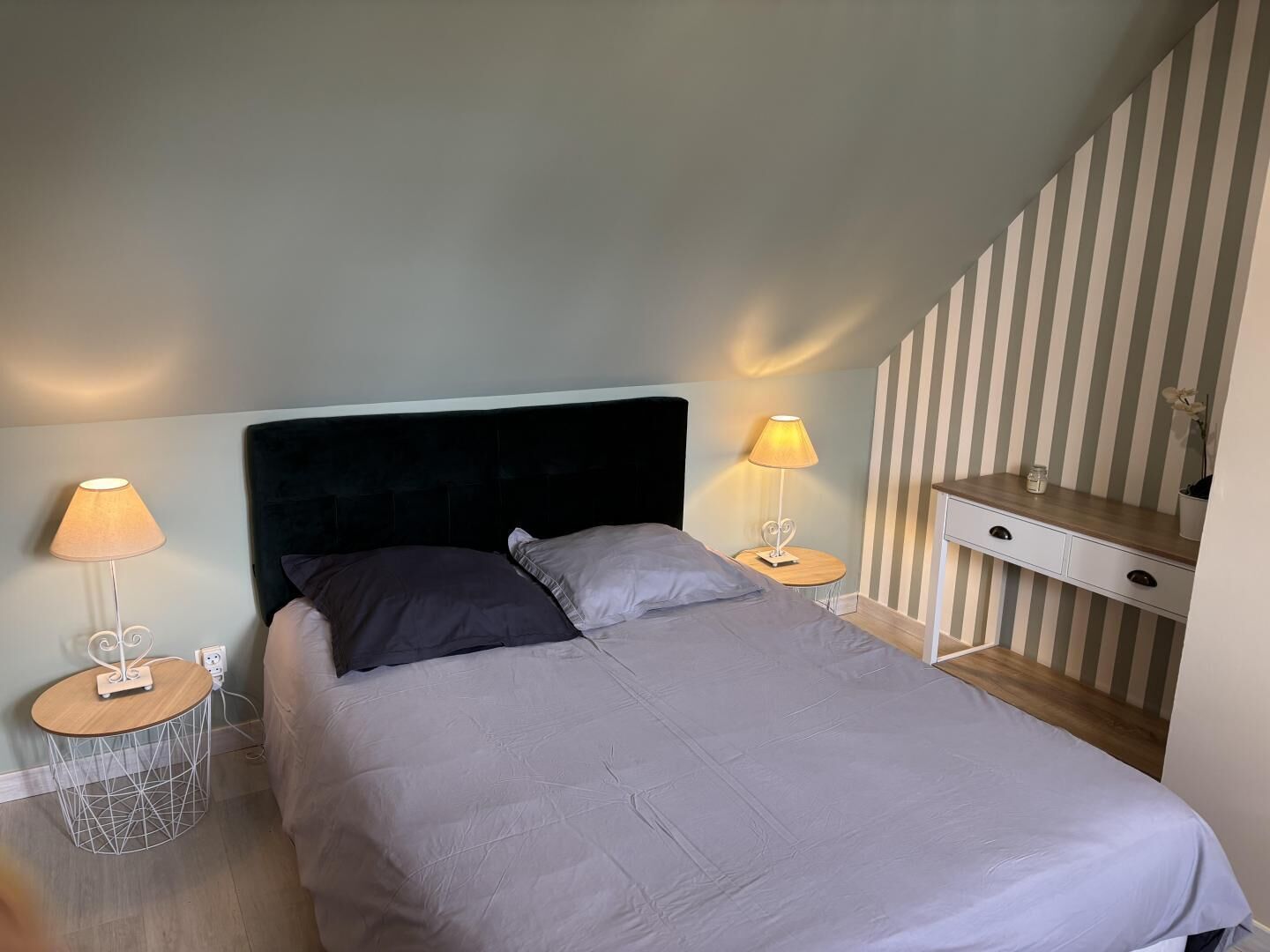 A bedroom with a bed, two bedside tables with lamps, a small round table, and a desk with drawers.