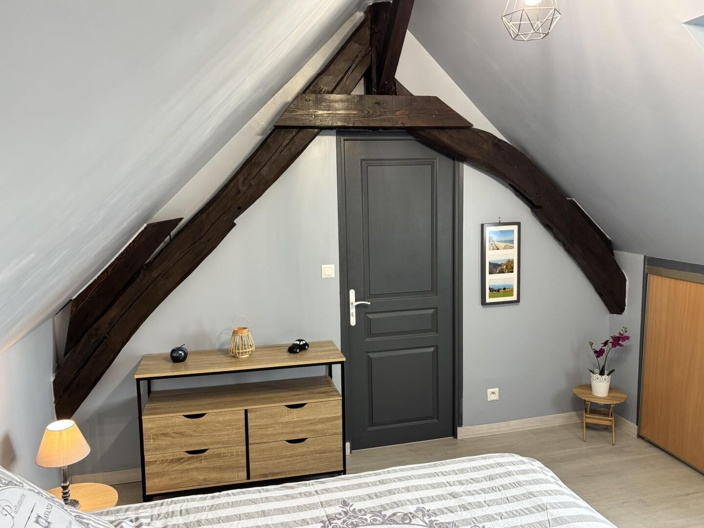 Ein Schlafzimmer mit schrägem Dach, einer dunklen Tür, einem kleinen Tisch mit Lampe sowie einem Bett mit gestreifter Bettdecke und einem gerahmten Bild an der Wand.