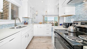 A modern kitchen with white cabinetry, a large island, a stove, and a view of the beach. - Incredible Oceanfront Home w/ Rooftop Deck & Cool, Coastal Interior! (Newport Beach)