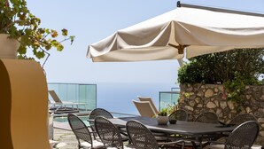 Un espace extérieur aménagé pour prendre les repas, avec une table et des chaises, un grand parasol et une vue sur la mer.