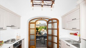 Une cuisine étroite avec des meubles blancs, un fourneau et une grande porte en bois donnant sur un patio extérieur.