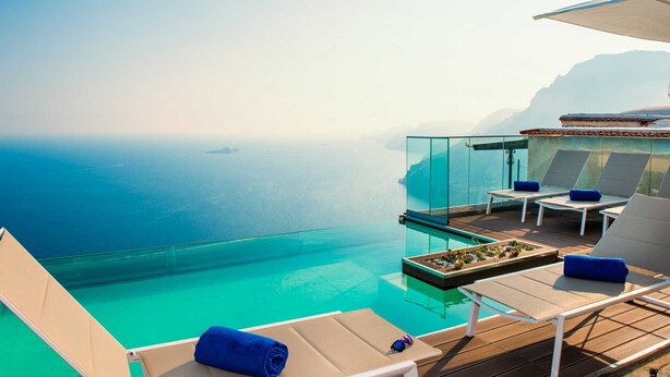 Une piscine à débordement luxueuse, avec une eau d’un bleu cristallin, offrant une vue imprenable sur une mer calme et des montagnes lointaines.