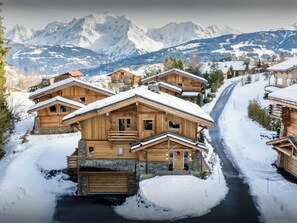 Een houten chalet met een stenen fundering, een garage en een oprit vol sneeuw.