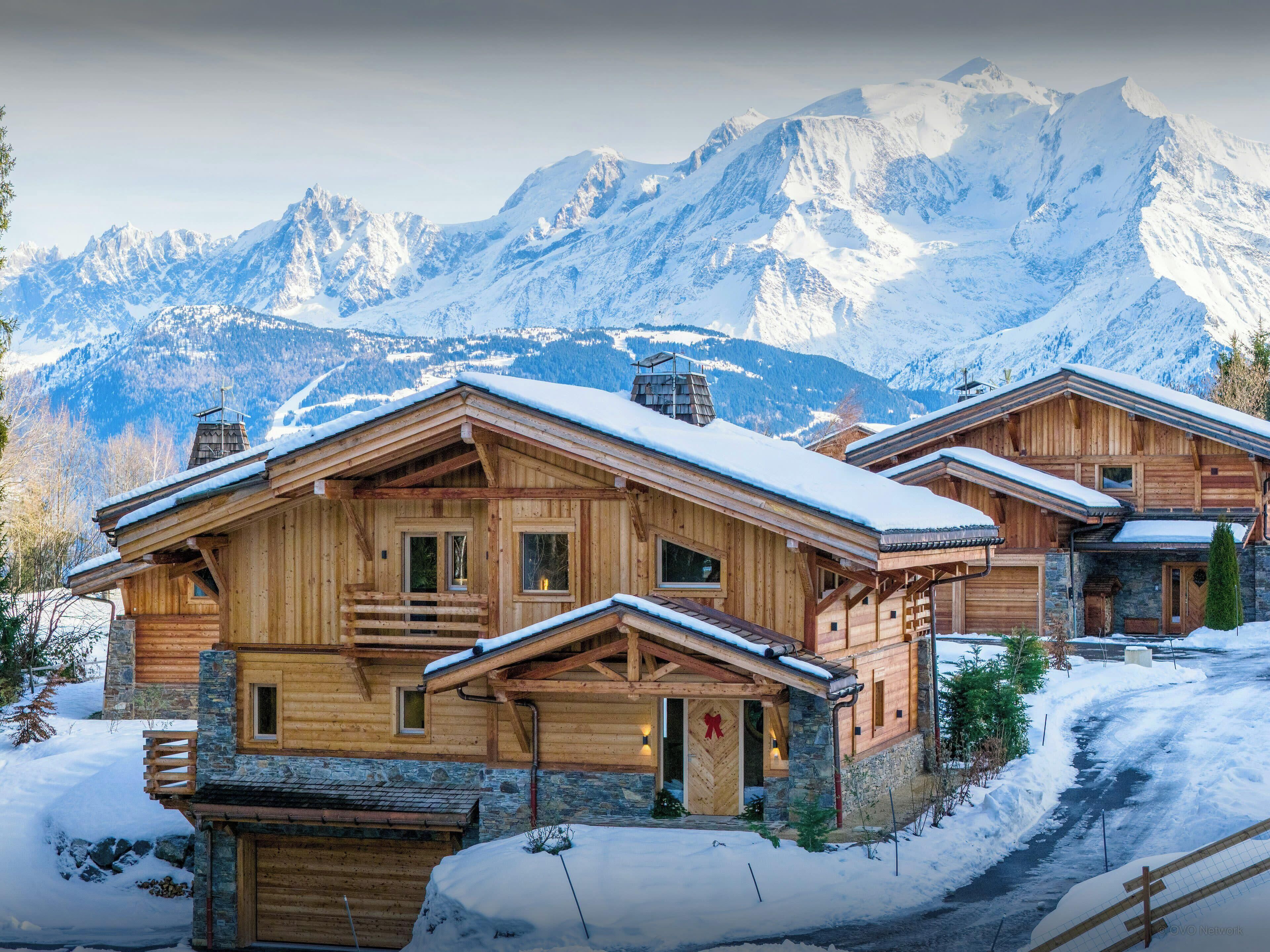 A wooden chalet with a stone foundation, set against a backdrop of snow-covered mountains.