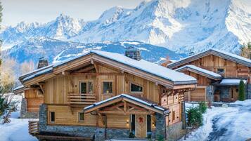 A wooden chalet with a stone foundation, set against a backdrop of snow-covered mountains.