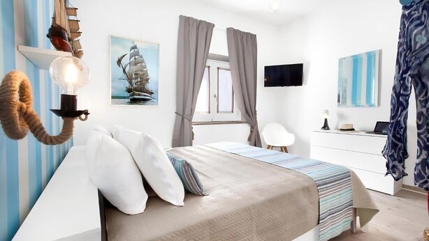 A bedroom with a bed, a desk, a chair, a ship painting, and a wall-mounted TV.