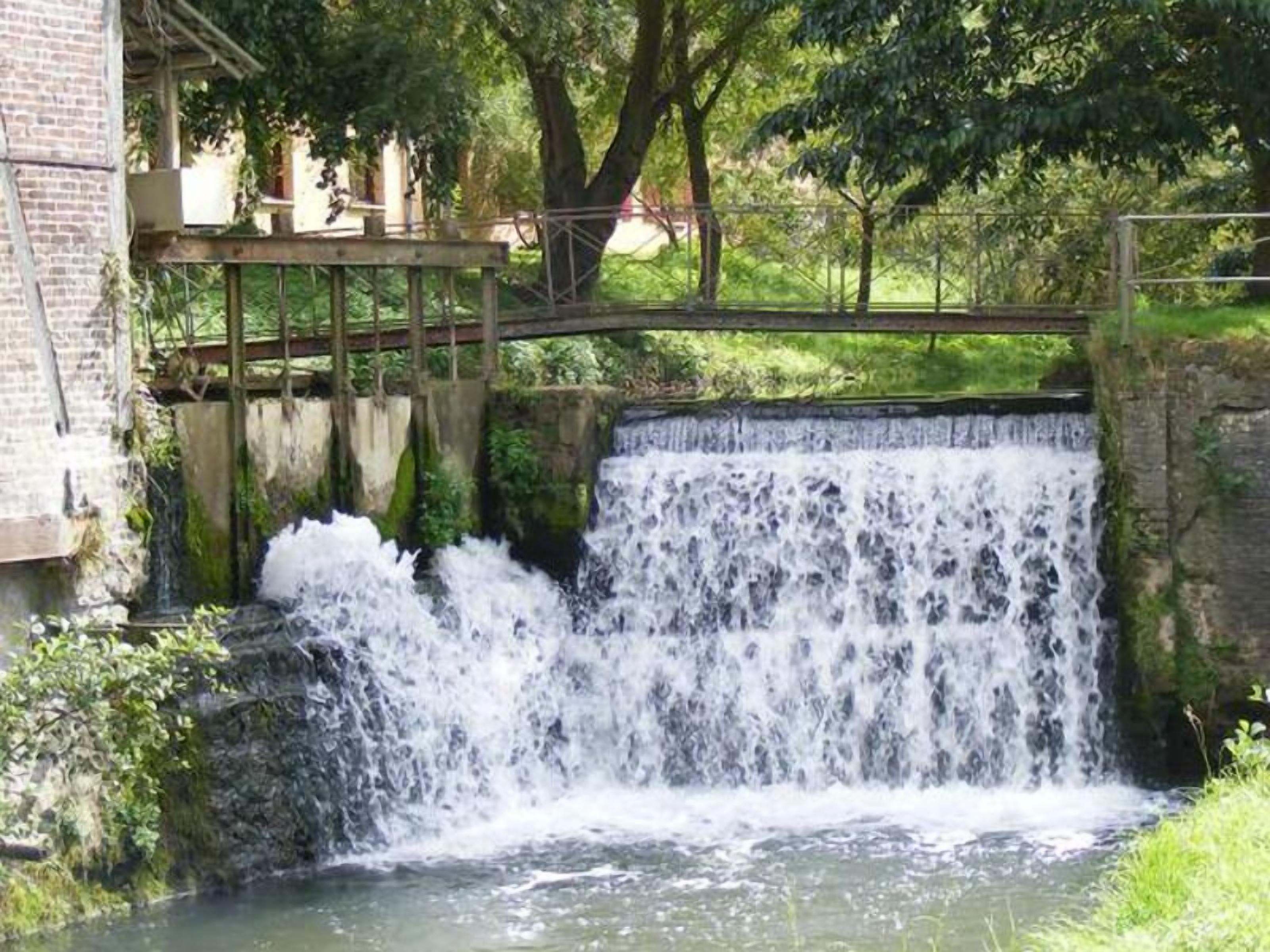 A small waterfall in a park with a wooden bridge and stone walls.