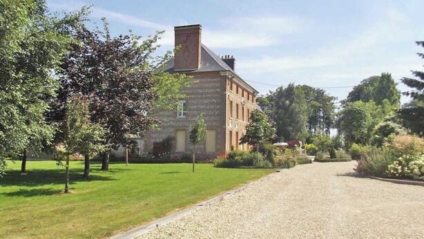 Un bâtiment historique à la façade en briques, entouré d’un jardin bien entretenu et bordé d’une allée en gravier.