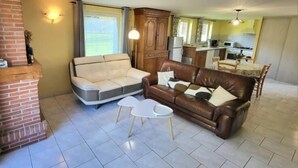 A living room with a sofa, a coffee table, and a fireplace. There is a kitchen in the background. - Gîtes de France® - L'Auge Normande (AVREMESNIL)