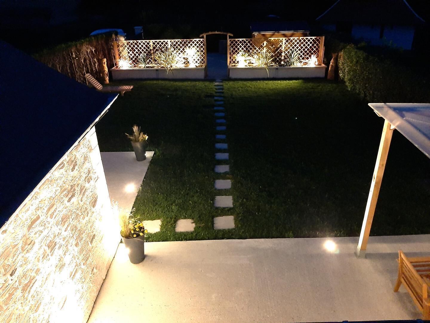 Een nachtzicht van een achtertuin met een verlicht pad, een houten terras en een kleine tuin.